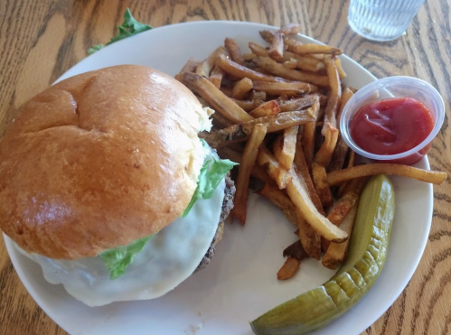 A plate with a cheeseburger topped with lettuce, fries, a pickle, and a small cup of ketchup.