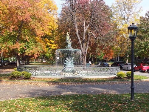 A decorative fountain surrounded by colorful autumn trees and fallen leaves, with parked cars in the background.