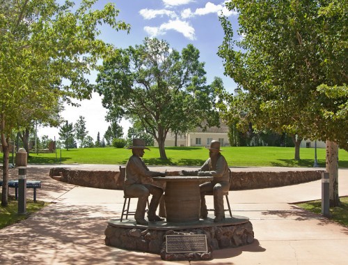 Bronze statue of two men sitting at a table in a park, surrounded by trees and a grassy area.