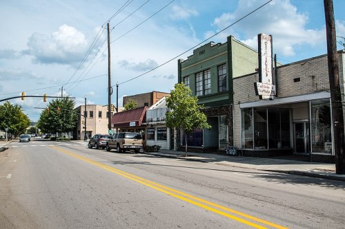 A quiet street scene featuring small businesses, parked cars, and a blue sky with scattered clouds.