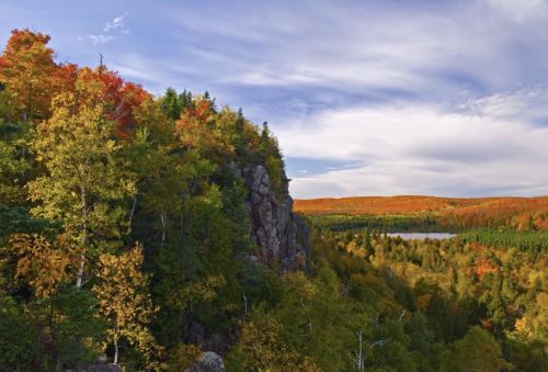 Vibrant autumn foliage on a rocky hillside overlooking a serene lake and distant forested hills under a blue sky.
