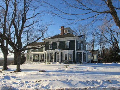 A large, green Victorian house surrounded by snow and bare trees under a clear blue sky.