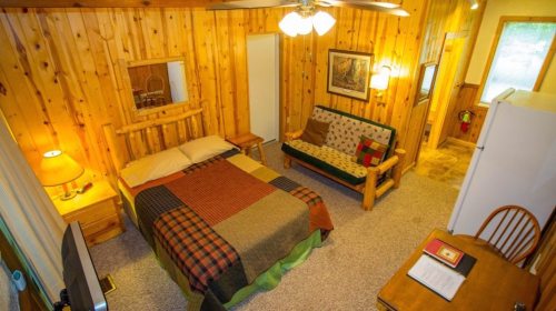 Cozy cabin interior featuring a wooden bed, futon, dining table, and warm wood paneling.
