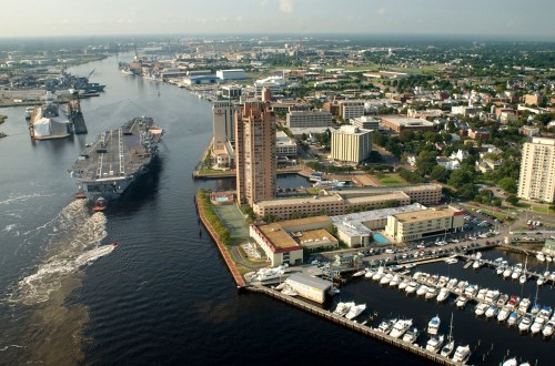 Aerial view of a city harbor with a naval ship, buildings, and boats along the waterfront.