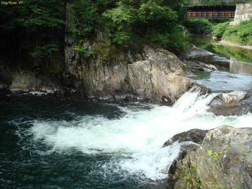 A serene river flows over rocks, surrounded by lush greenery and a bridge in the background.
