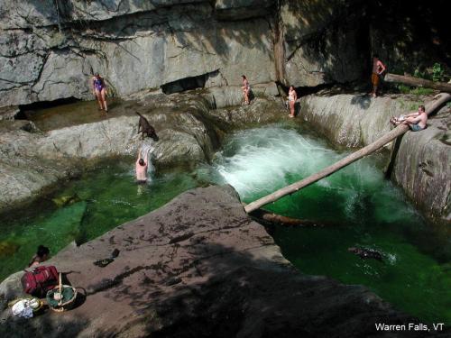 A scenic view of Warren Falls, VT, with people swimming and relaxing in a natural pool surrounded by rocks and trees.