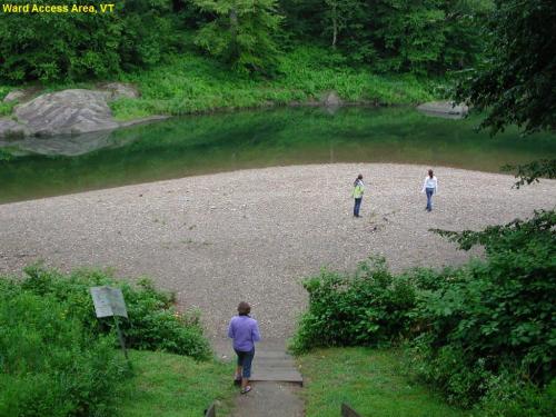 Two people walking on a sandy area by a calm river, surrounded by greenery and rocks, with a third person in the foreground.