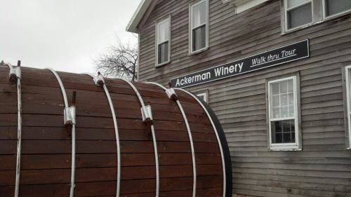 A large wooden wine barrel in front of the Ackerman Winery building with a sign for a walk-thru tour.