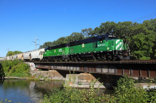 A green and black train crosses a bridge over a river, surrounded by trees and a clear blue sky.