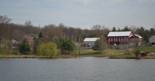A serene lakeside view featuring a red barn, white buildings, and lush greenery under a cloudy sky.