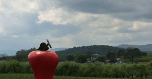 A large red apple sculpture in a green landscape with rolling hills and cloudy skies in the background.