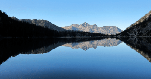 A serene lake reflects mountains and a clear blue sky, surrounded by evergreen trees.