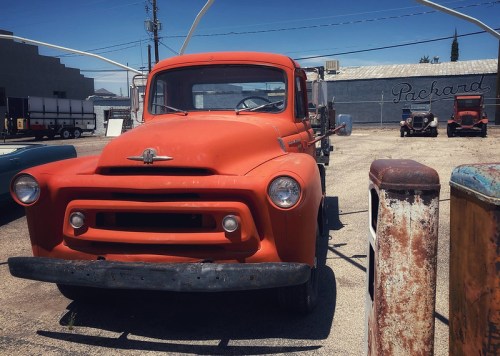 An orange vintage truck parked in a lot, with a faded building and other old vehicles in the background.