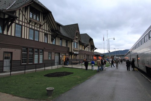 Historic train station with a distinctive architectural style, people gathered outside, and a train parked nearby.