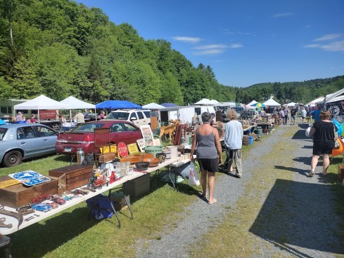 A bustling outdoor market with vendors selling antiques and crafts, surrounded by green trees and blue skies.
