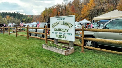 Sign for an antique and flea market in Wilmington, with tents and cars in the background, surrounded by autumn foliage.