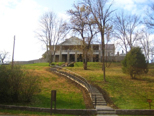 A large, two-story house on a grassy hill, with a winding stone staircase leading up to it and trees surrounding the area.