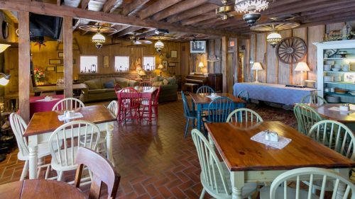 Cozy restaurant interior with wooden beams, colorful chairs, and tables, featuring a piano and warm lighting.