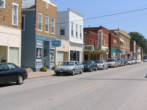 A quaint street lined with small shops and parked cars under a clear blue sky.