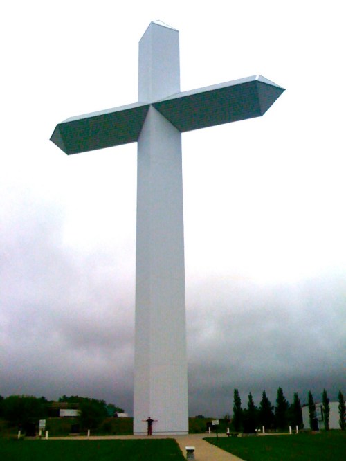 A tall, white cross stands against a cloudy sky, surrounded by green grass and trees.