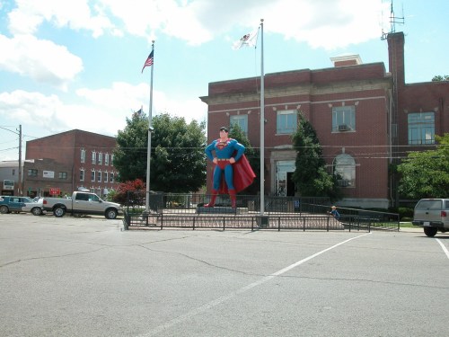 A large Superman statue stands in front of a brick building, surrounded by flags and parked cars in a small town.