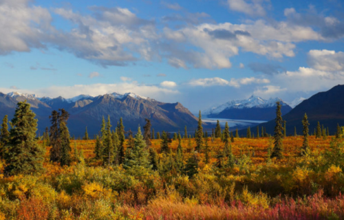 A scenic landscape featuring mountains, a lake, and colorful autumn foliage under a partly cloudy sky.