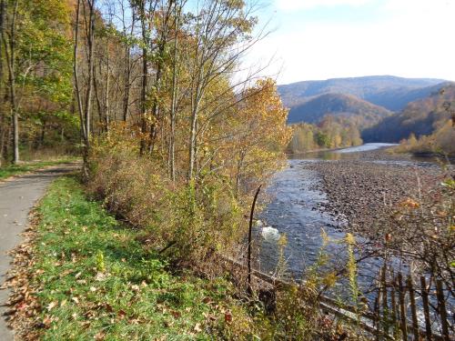 A scenic riverbank surrounded by autumn trees, with a winding path alongside the water and mountains in the background.