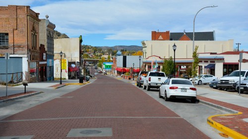 A quiet street lined with shops and parked cars, featuring brick pavement and a clear blue sky.