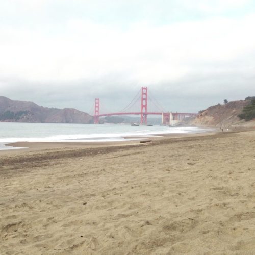 A sandy beach with gentle waves and the Golden Gate Bridge in the background under a cloudy sky.