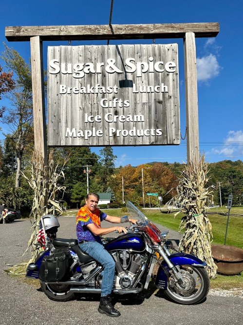 A person in a tie-dye shirt sits on a motorcycle in front of a sign for "Sugar & Spice" restaurant and shop.