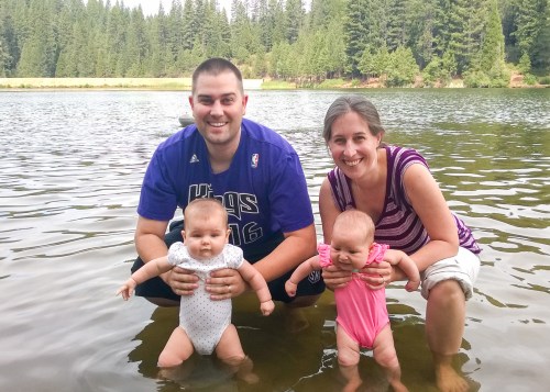 A man and woman hold two babies in shallow water, surrounded by trees and a calm lake. Everyone is smiling.
