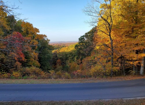 A scenic view of colorful autumn trees overlooking a valley and a winding road under a clear blue sky.