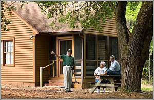 Three people sit outside a wooden house, one gesturing while the others listen, surrounded by trees.