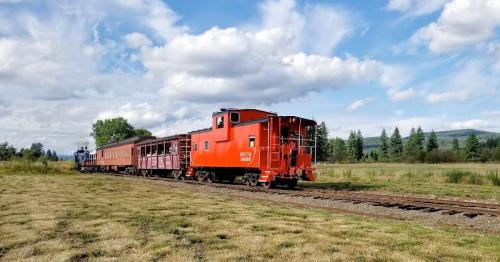 A red caboose and vintage train cars on a grassy railway under a blue sky with fluffy clouds.
