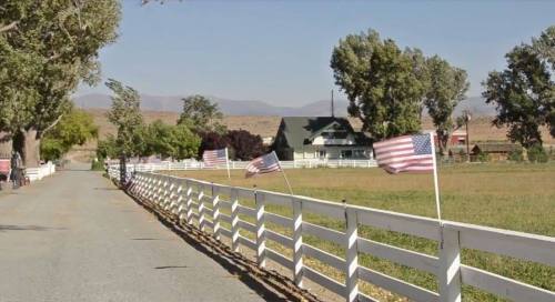 A rural road lined with white fences and American flags, leading to a house surrounded by trees and mountains in the background.