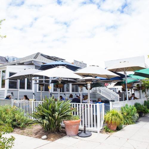 A cozy restaurant patio with umbrellas, plants, and diners enjoying their meals on a sunny day.