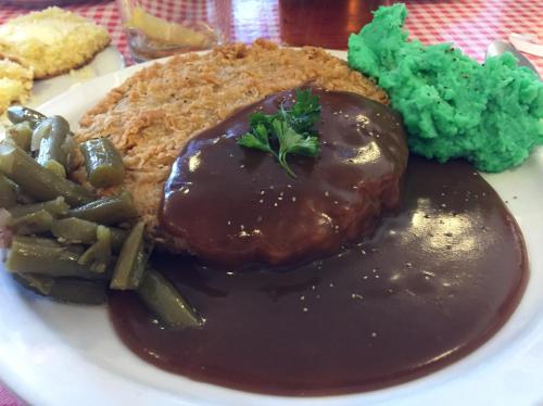 A plate of food featuring green mashed potatoes, green beans, and a breaded meat patty topped with brown gravy.
