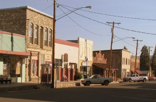 A quiet street scene featuring historic buildings and parked cars in a small town. Power lines stretch overhead.