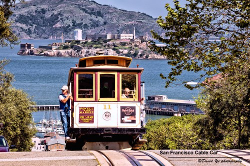A San Francisco cable car travels down a hill with Alcatraz Island visible in the background and a clear blue sky.