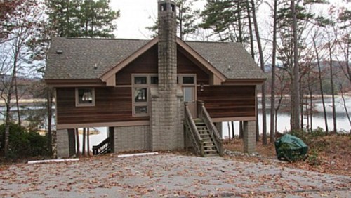A modern house with a sloped roof, wooden siding, and a stone chimney, surrounded by trees and near a lake.