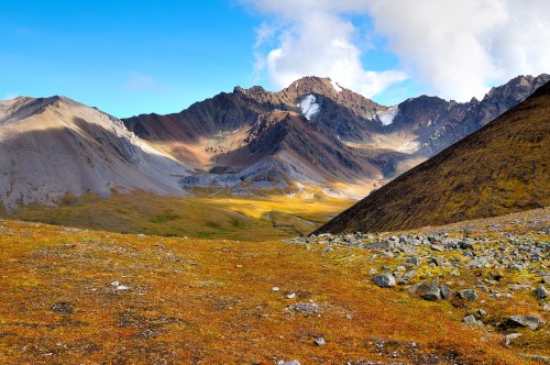 A scenic mountain landscape with colorful autumn foliage and rocky terrain under a blue sky with scattered clouds.