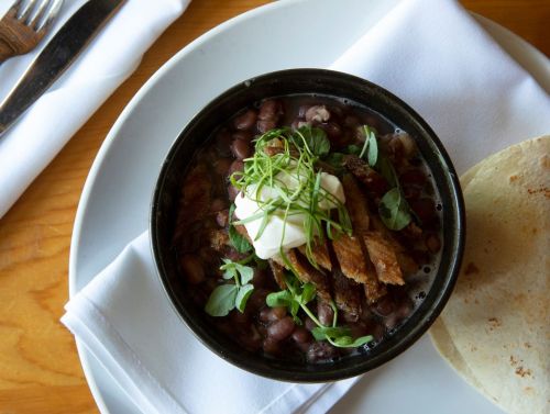 A bowl of black beans topped with crispy meat, sour cream, and herbs, served with a side of tortillas on a plate.