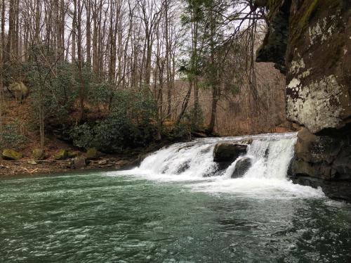 A serene waterfall cascades into a clear green pool, surrounded by trees and rocky cliffs.