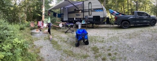 A campsite scene featuring a camper, truck, chairs, and a child playing near a fire pit in a wooded area.