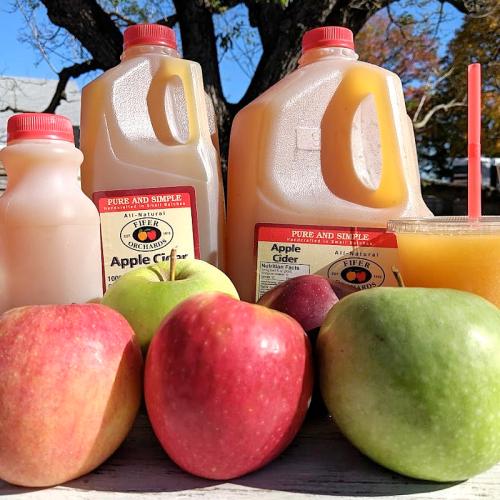 Two jugs of apple cider, a cup of cider, and various apples on a table with a tree in the background.