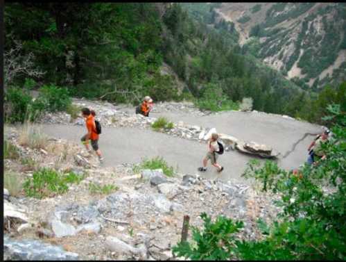 Hikers ascend a rocky trail surrounded by trees and mountains, with a winding path visible in the background.