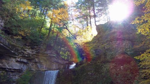 A waterfall cascades down rocky cliffs, surrounded by vibrant autumn foliage and sunlight creating a rainbow effect.