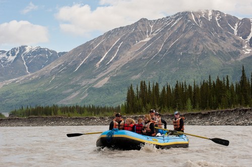 A group of people rafting on a river with mountains and trees in the background under a partly cloudy sky.
