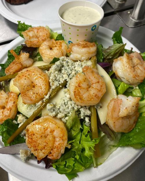 A fresh salad topped with grilled shrimp, blue cheese, and a side of ranch dressing in a cup.