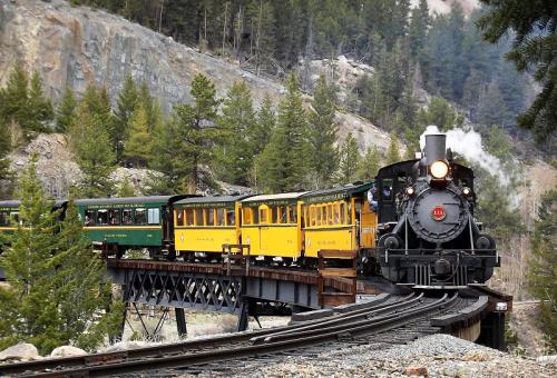 A vintage steam train crosses a wooden bridge surrounded by lush green trees and rocky terrain.